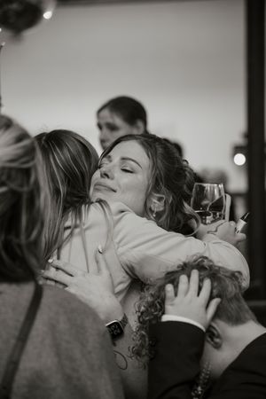 Emotional black and white photo of woman hugging guests while holding wine glass at wedding.