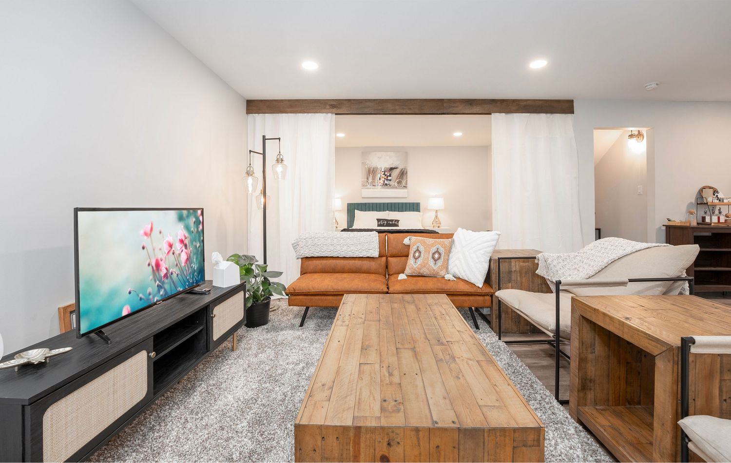Modern studio apartment with rustic wood coffee table, flat-screen TV, and sheer curtain bedroom divider.