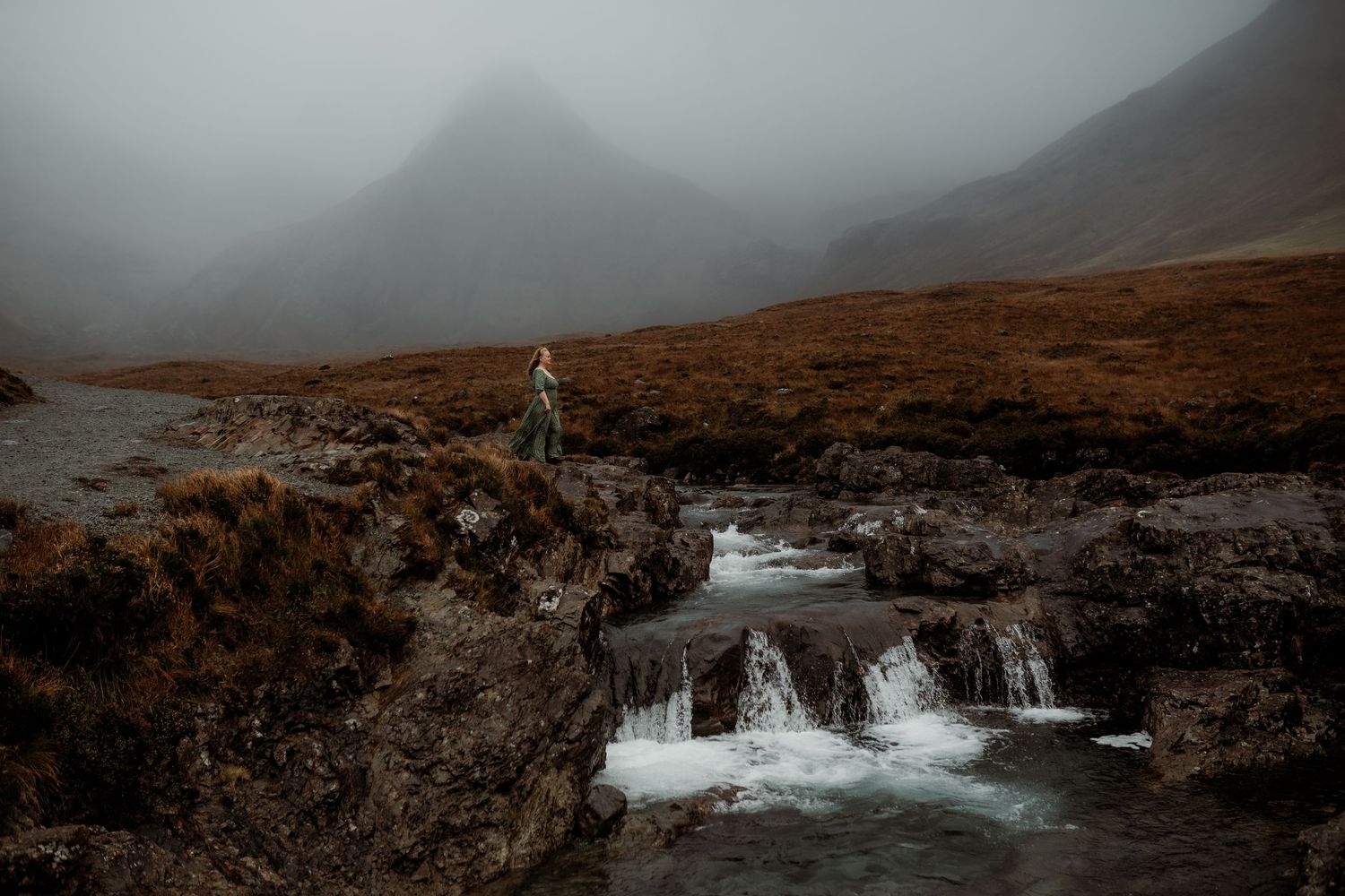 Person standing on rocky terrain beside cascading waterfalls in a misty Scottish Highland valley with mountains in the background.