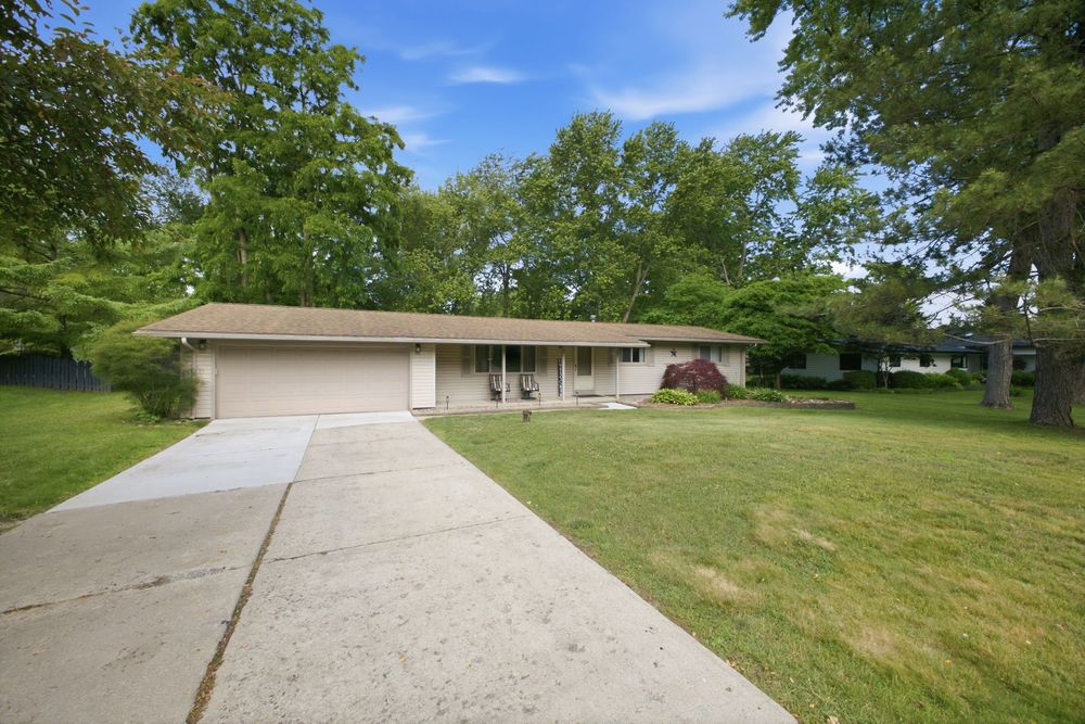 Single-story ranch home with attached garage, long concrete driveway, and lush green lawn shaded by mature trees.