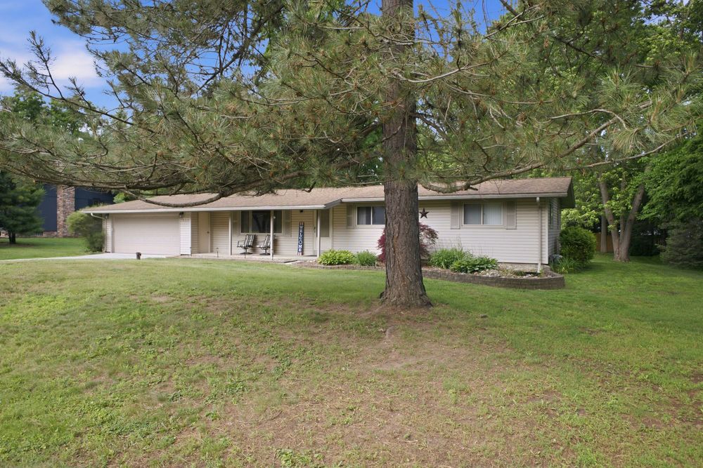 Ranch-style home surrounded by mature pine trees with a well-maintained green lawn in a peaceful suburban setting.