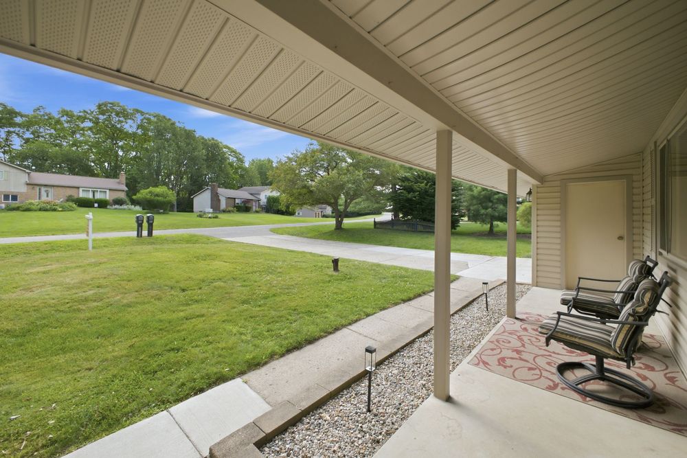 Covered front porch with rocking chairs overlooks a lush green lawn and quiet suburban neighborhood on a sunny day.
