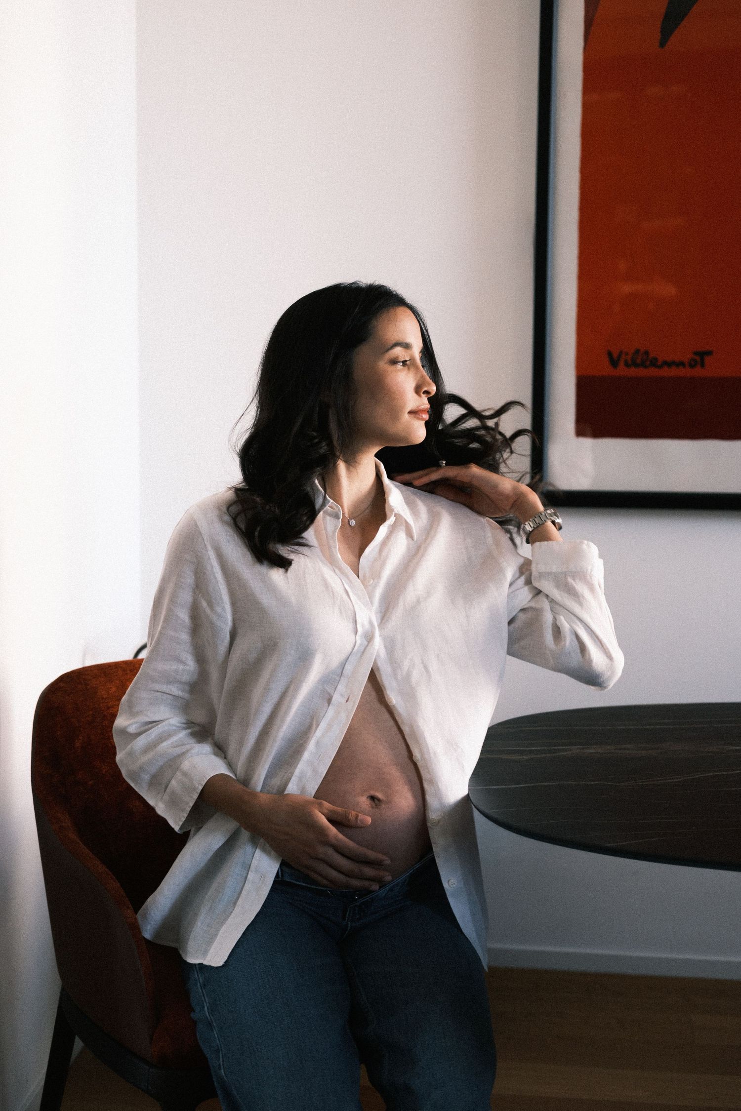 Pregnant woman in white shirt and jeans cradling baby bump, gazing sideways in modern room with artwork.