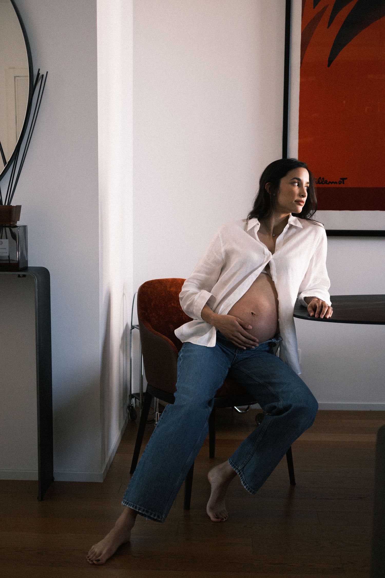 Pregnant woman in white shirt and jeans sitting on chair, cradling baby bump, gazing thoughtfully to the side.
