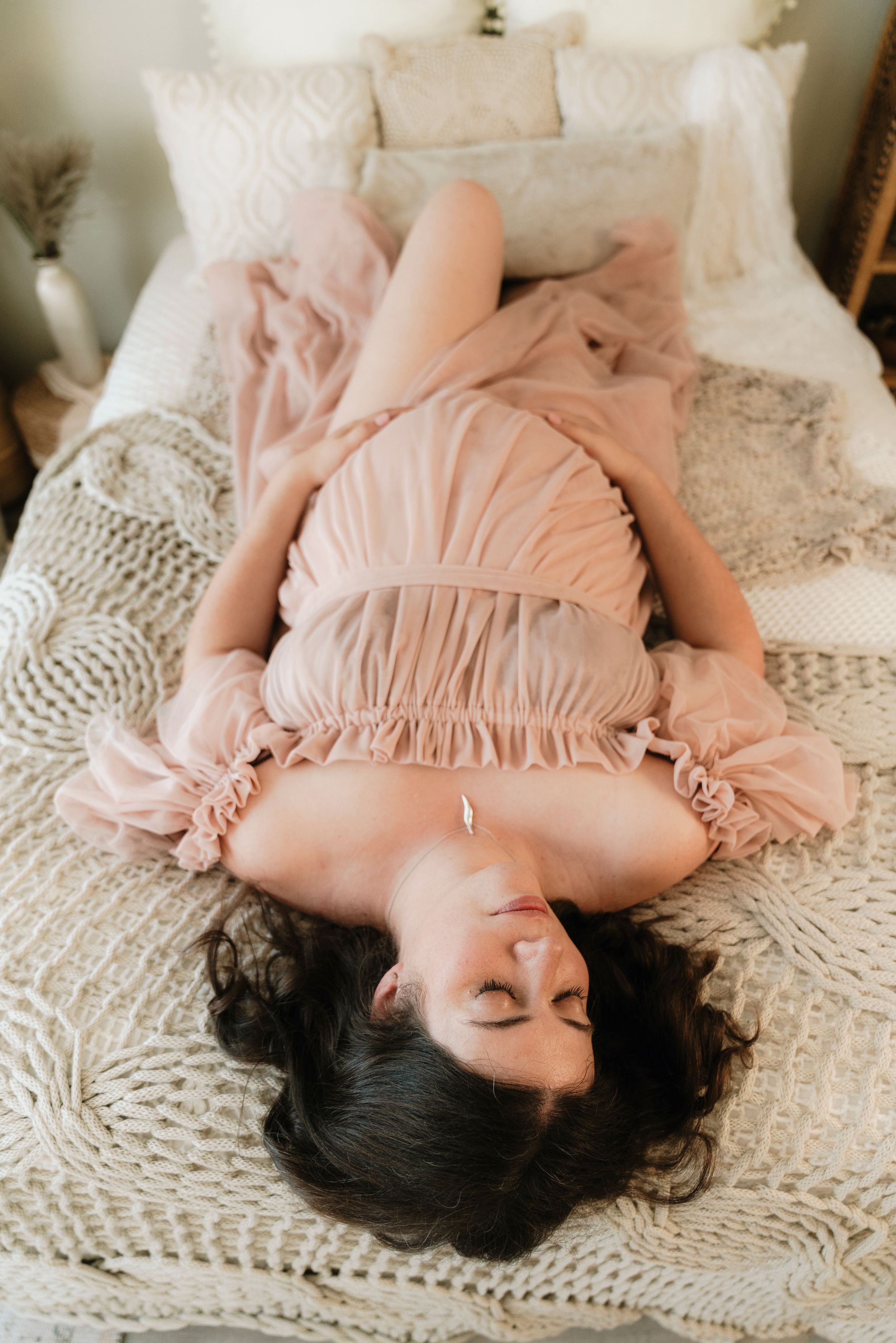 A woman in a blush pink ruffled dress lies gracefully on a white knit blanket on a bed.