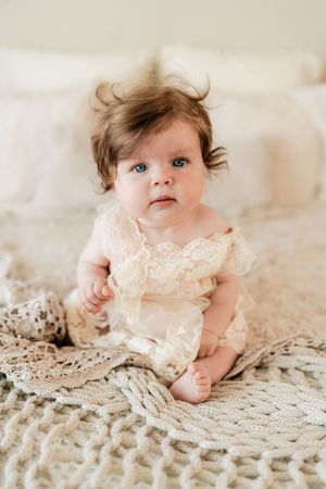 Adorable baby girl with blue eyes sitting on a knit blanket wearing a cream lace romper dress.