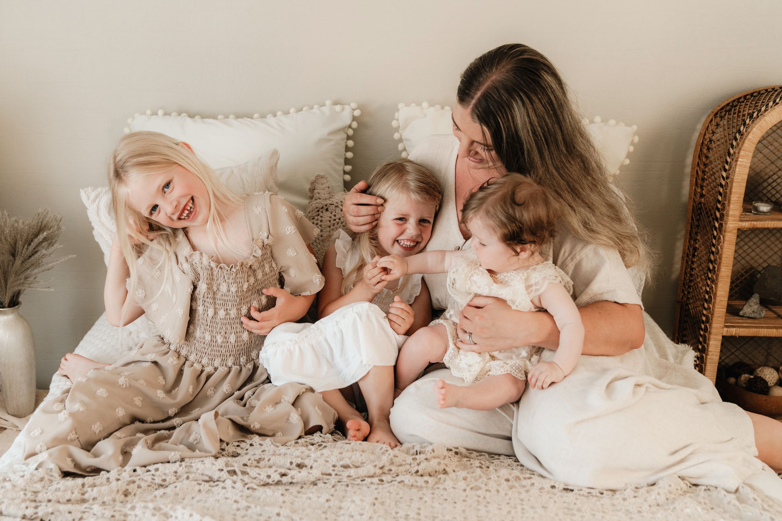A mother smiles warmly with her three young daughters sitting together on a cozy white bed.