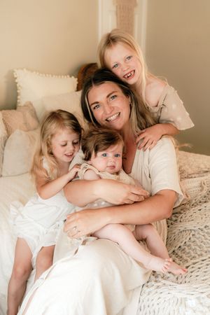 A smiling mother sits on a bed with her three young children in a warm, neutral-toned room.
