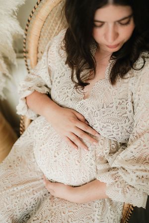 Pregnant woman in white lace dress cradling baby bump while seated in rattan chair.