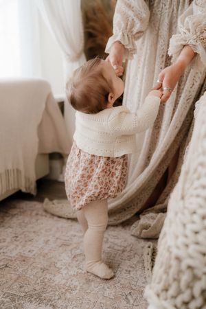 A baby in a floral romper and white knit cardigan takes first steps, holding an adult's hands in a bright room.