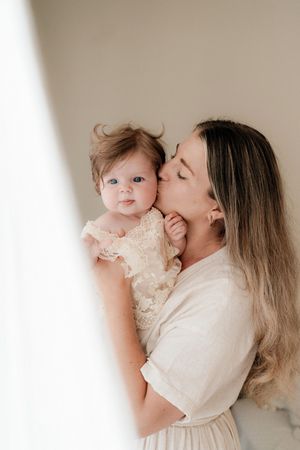 A loving mother gently kisses her baby girl's cheek while holding her in a bright, neutral-toned room.