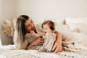 A young mother and baby girl laugh together on a cozy white knit blanket in a bright, minimalist bedroom.