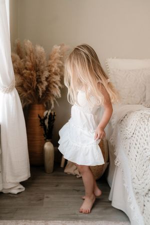 Young blonde girl in white ruffle dress dancing barefoot in boho bedroom with pampas grass decor.