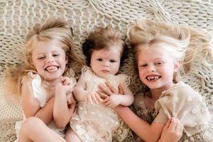 Three young sisters in cream dresses lying together on a knit blanket, smiling and laughing joyfully.