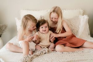 Three young blonde girls sit on a white bed, smiling and laughing together in soft natural light.