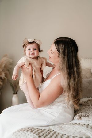 A young mother in a white dress holds her smiling baby girl in a beige outfit on a cozy bed.