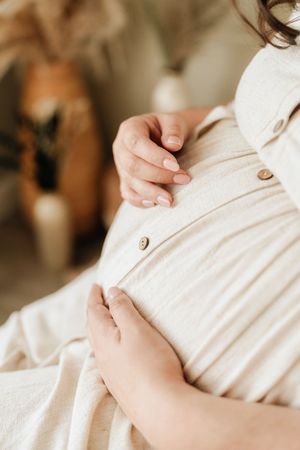Pregnant woman in beige linen dress gently cradling her baby bump with both hands in a warm, softly lit room.