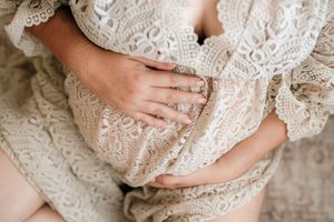 Pregnant woman in lace dress cradling baby bump with ringed hands in elegant maternity photo.