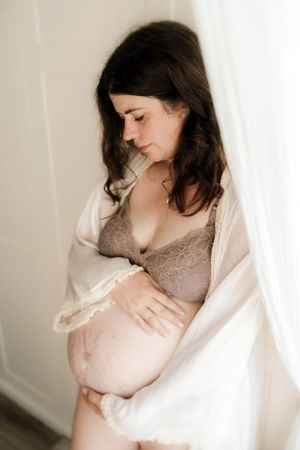 Pregnant woman in lace bra and sheer robe cradling her baby bump by a bright window.