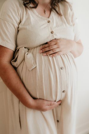 Pregnant woman in a beige linen button-down dress gently cradling her baby bump with both hands.