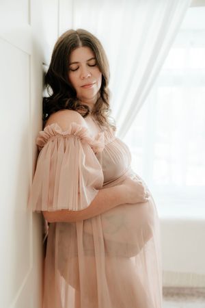 Pregnant woman in blush off-shoulder tulle gown cradling her baby bump, leaning against white wall in soft natural light.