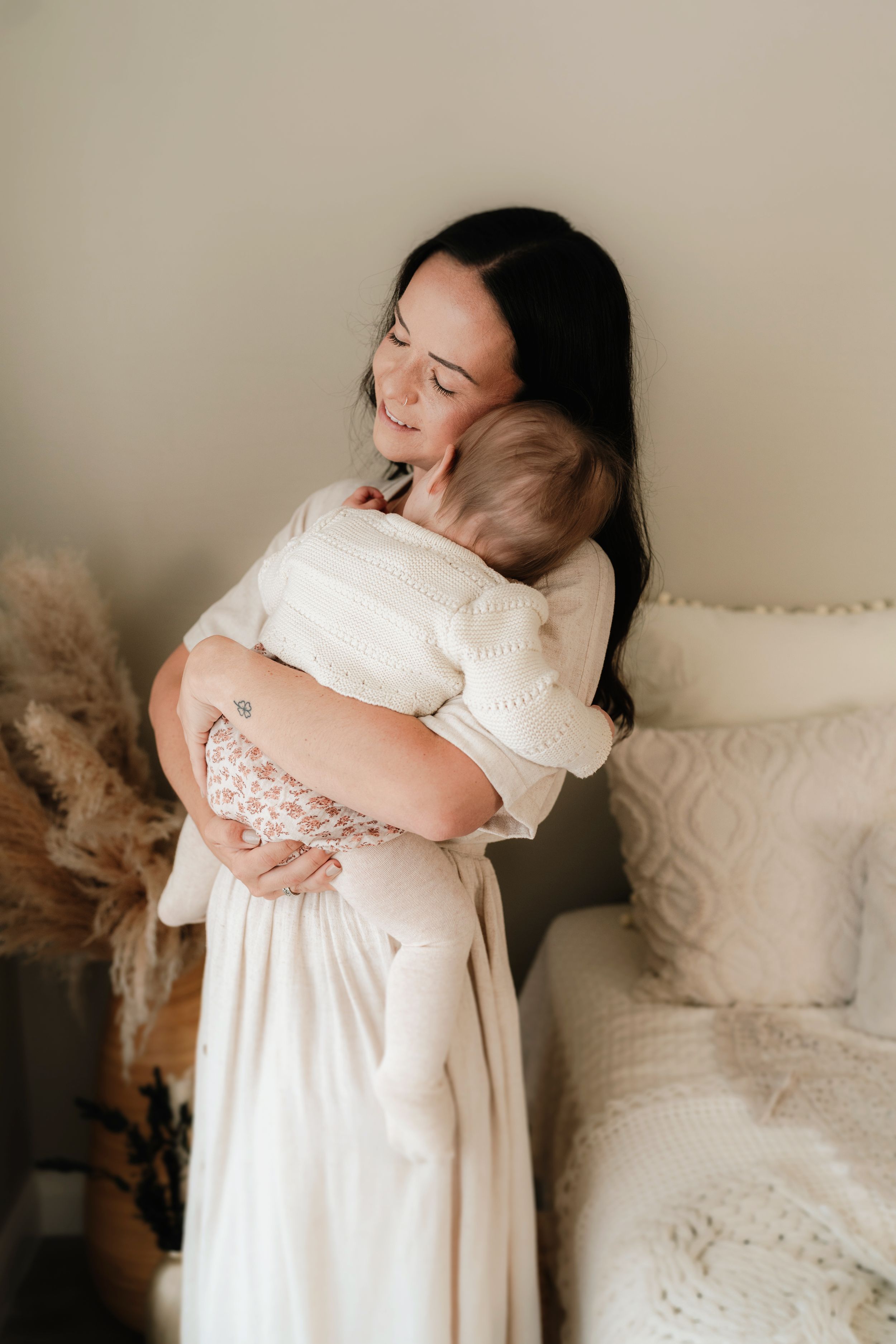 A dark-haired mother in a white dress tenderly holds her baby in a softly lit, cozy bedroom setting.