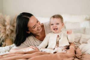 A smiling mother with dark hair lies on a bed with her laughing baby dressed in a cream cardigan.