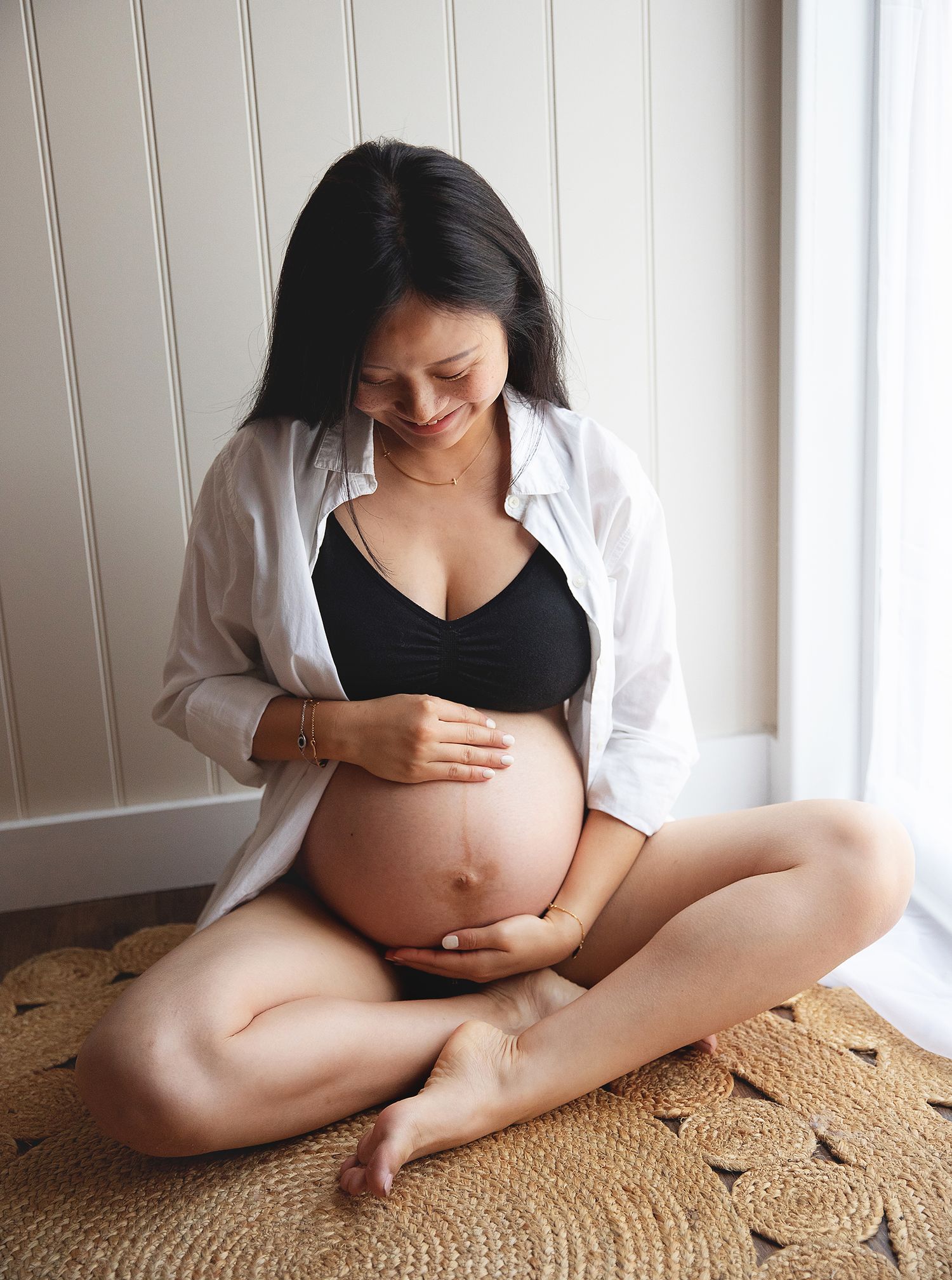 Pregnant woman sitting on a rug, holding her baby bump, wearing a black bralette and open white shirt. During a photoshoot in Bexley