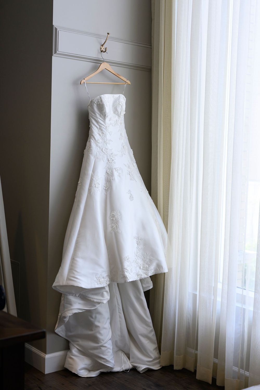 Elegant strapless white wedding dress with lace details hanging on a wooden hanger near a bright window.
