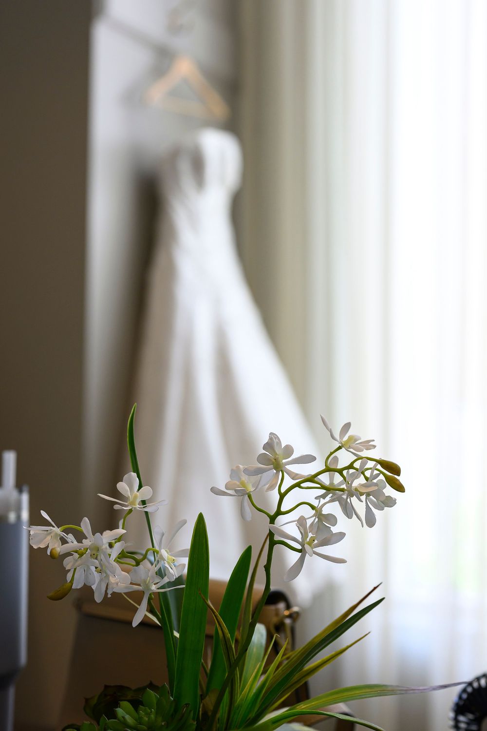 White wedding dress hanging on hanger with elegant white orchid flowers in foreground, bridal preparation scene.