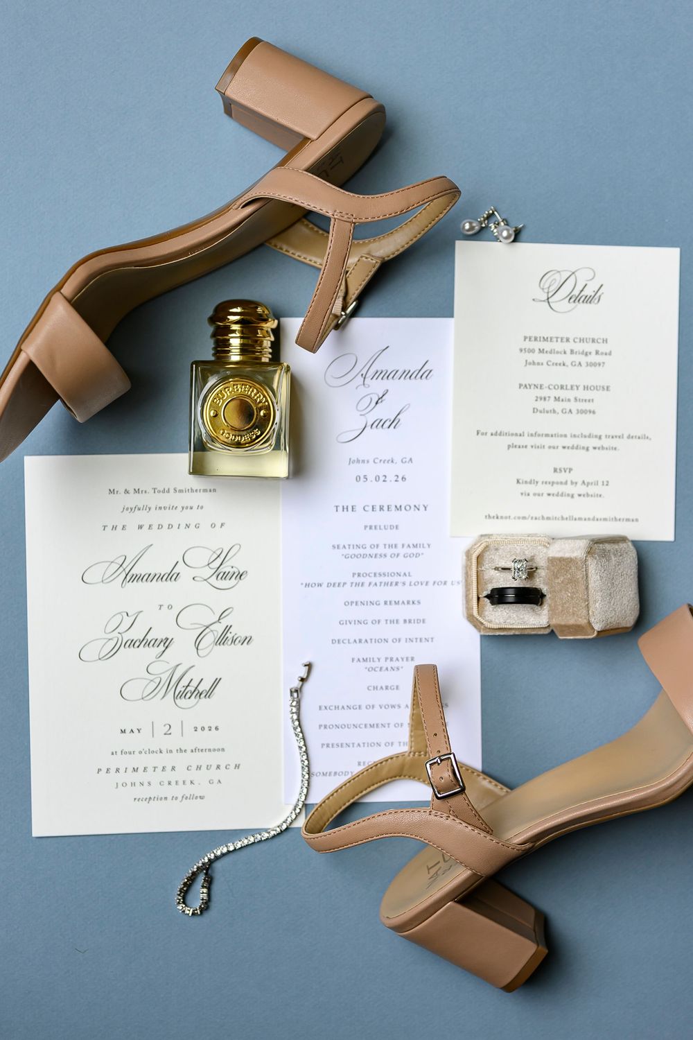 Elegant wedding flat lay featuring nude heels, perfume bottle, invitation suite, diamond ring box, and bracelet on blue background.
