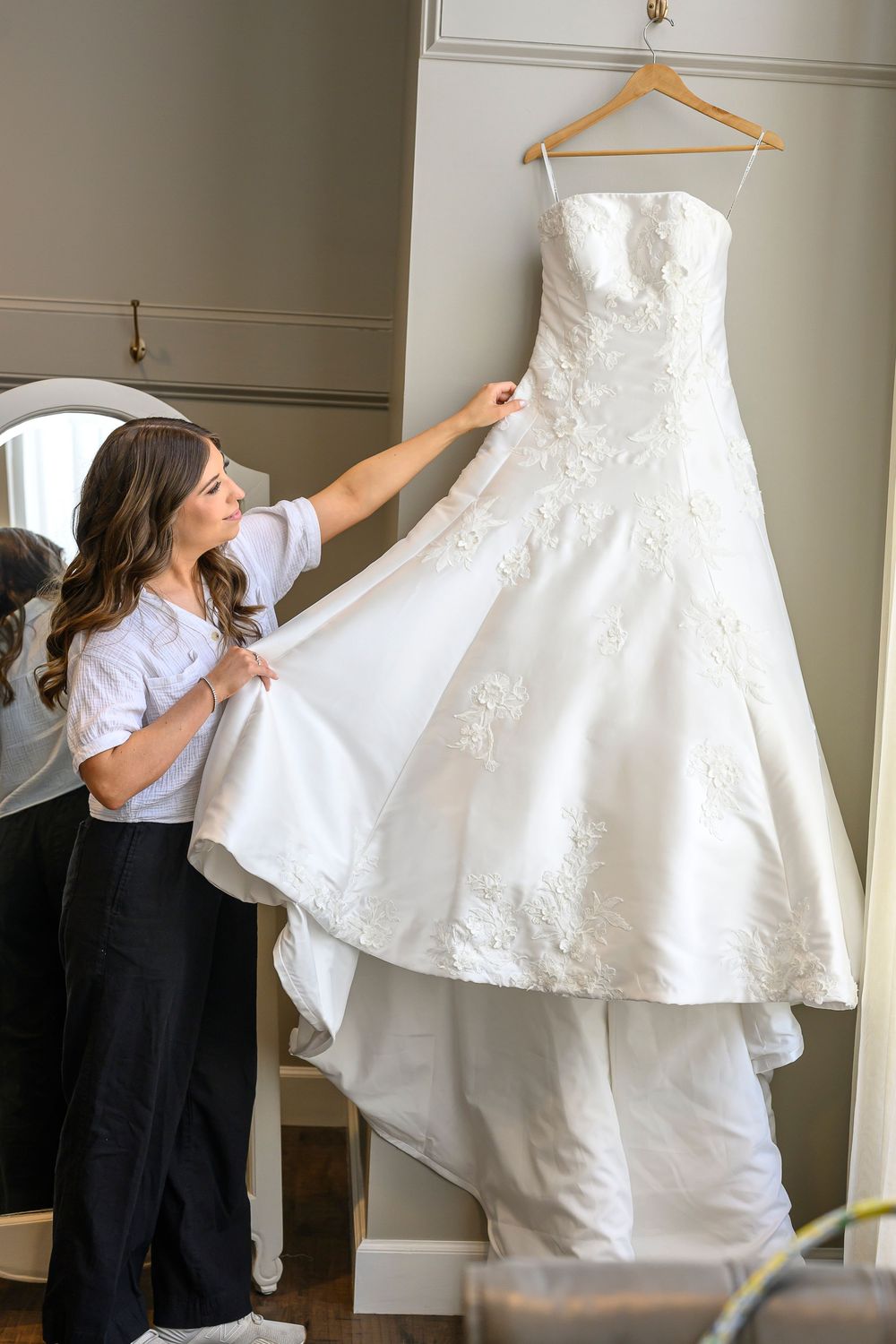 Bride admiring her white lace A-line wedding dress hanging on a wooden hanger in a bridal suite.