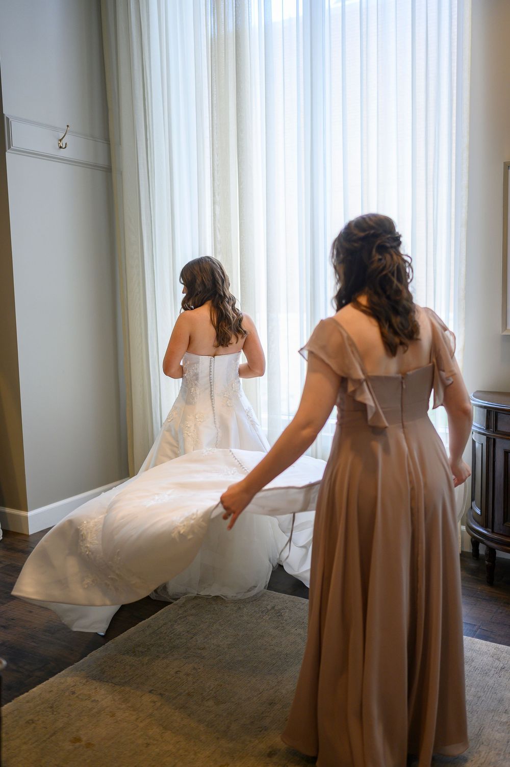 Bride in white gown with bridesmaid in taupe dress holding her train near bright windows before wedding ceremony.