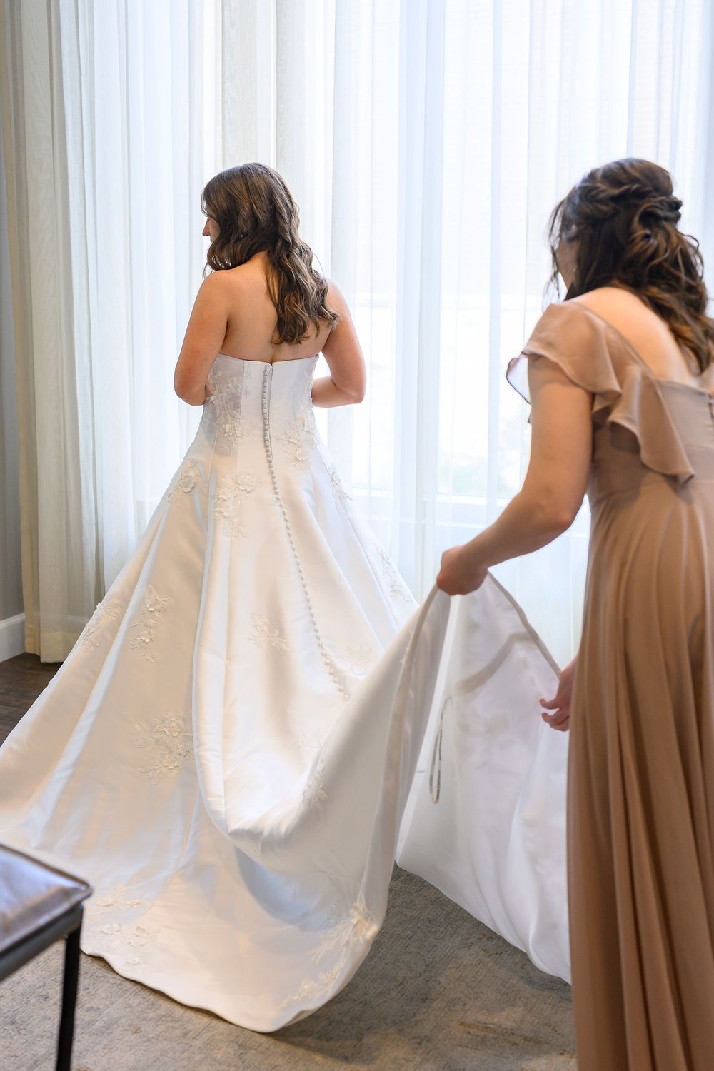 Bridesmaid in taupe dress helps bride in strapless white ball gown with floral details near bright window.