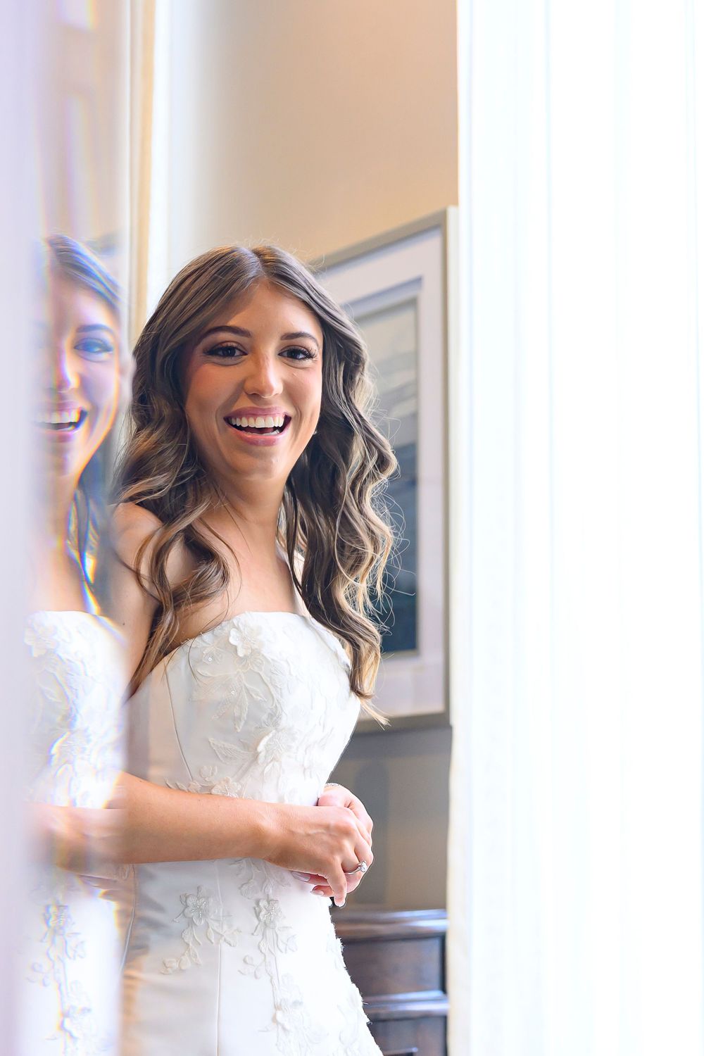 Smiling bride in white lace wedding dress standing near a mirror, with wavy hair and radiant makeup.