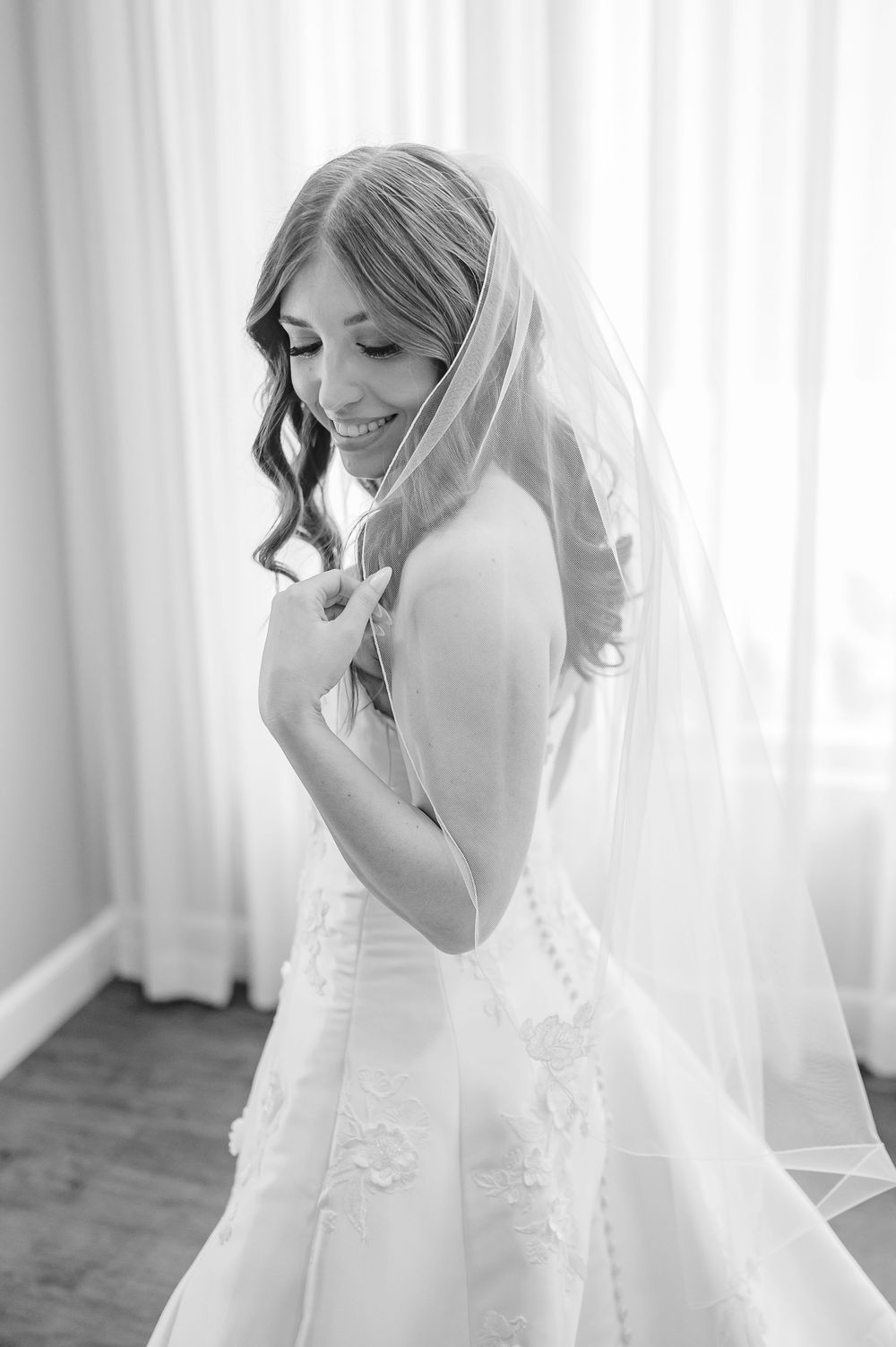 Black and white photo of a smiling bride in a lace wedding gown and veil, posing gracefully near a bright window.