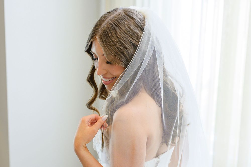 Smiling bride in a white wedding dress and veil looking down while getting ready near a bright window.
