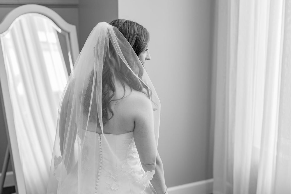 Black and white photo of a bride in a strapless gown and veil, standing by a window in soft natural light.