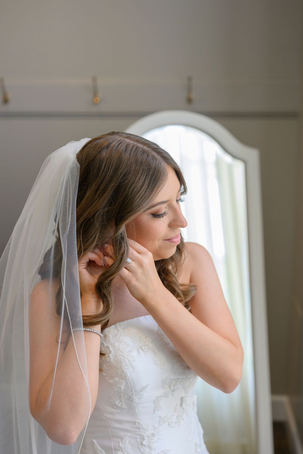 Bride in white lace gown and veil putting on earrings before her wedding ceremony near an arched mirror.