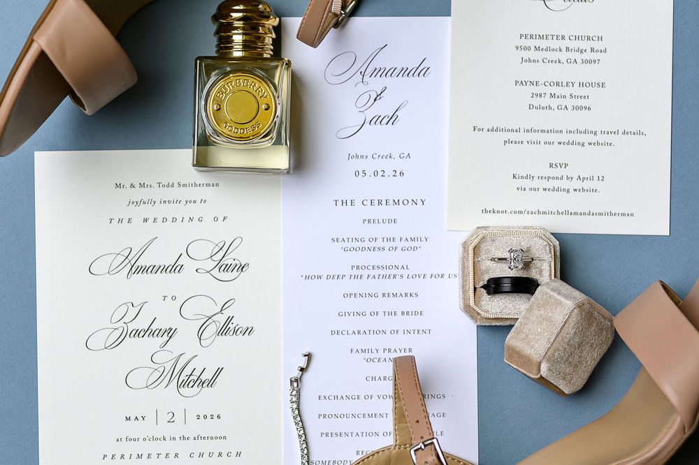 Elegant wedding invitation suite with calligraphy, perfume bottle, rings, and nude heels on a blue gray background.