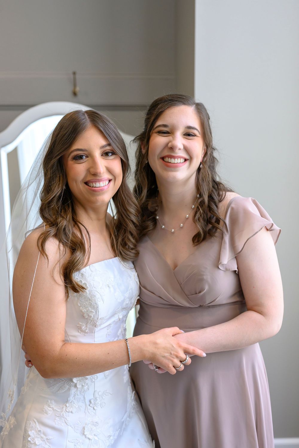 Bride in white lace gown and veil smiles with bridesmaid in taupe satin dress at wedding celebration.