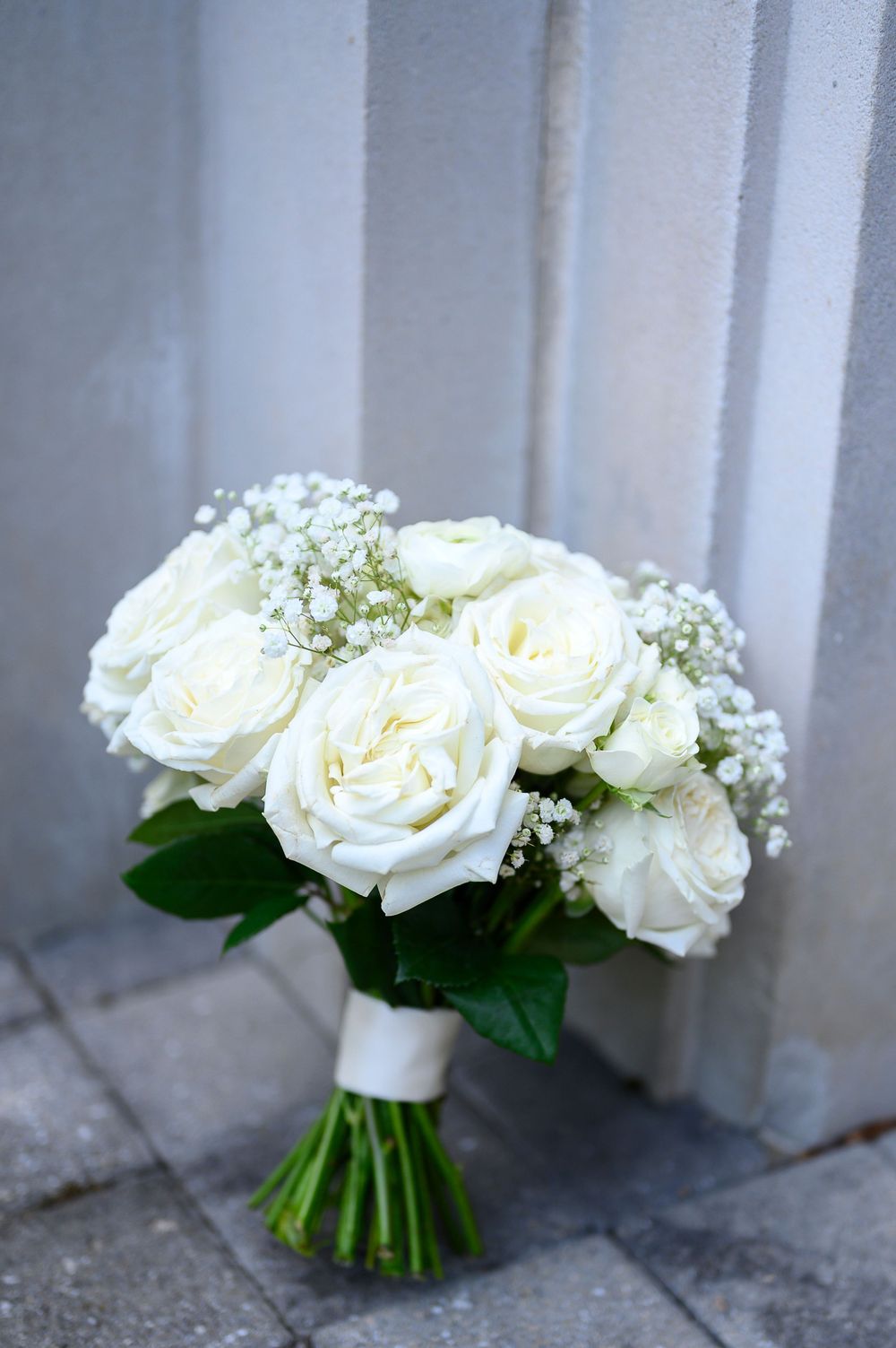Elegant bridal bouquet of white roses and baby's breath tied with white ribbon, leaning against a gray wall.