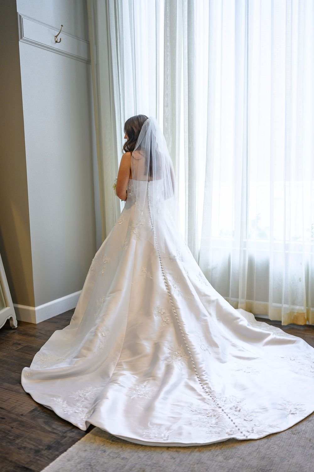 Bride in elegant white wedding gown with long train and veil stands by a sunlit window on her wedding day.