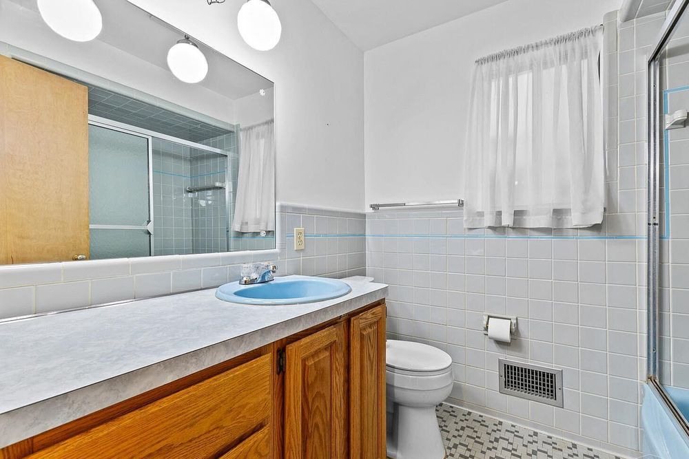 Bathroom with wood vanity, blue sink, large mirror, tiled walls, toilet, and glass shower enclosure.