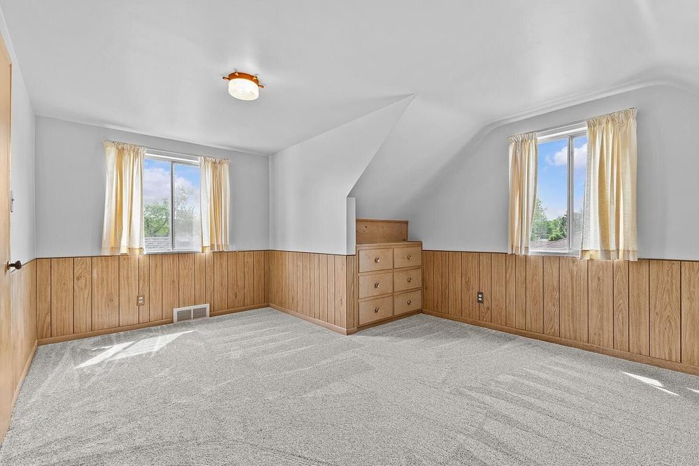 Spacious bedroom with vaulted ceiling, wood paneling, built-in dresser, carpet, and two windows.