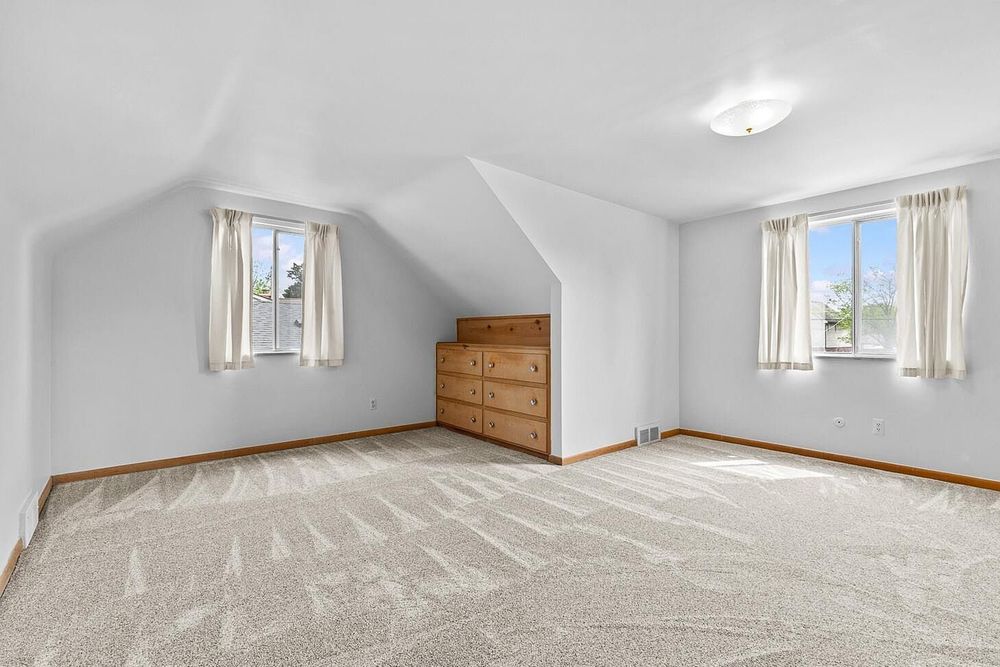 Bright attic bonus room with vaulted ceiling, neutral carpet, built-in dresser, and two windows with curtains.