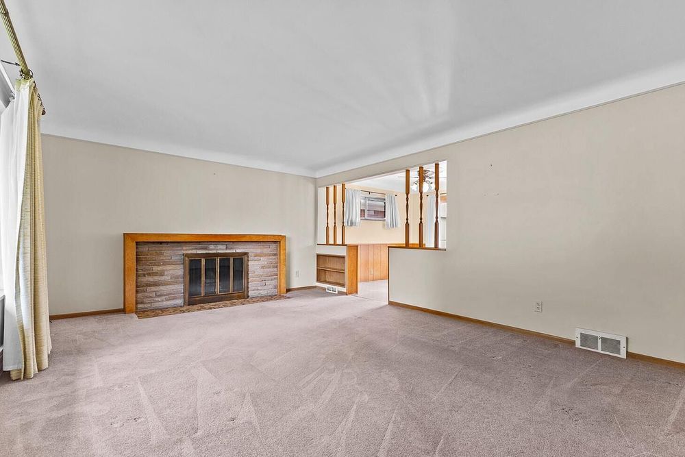 Empty living room with beige carpet, wood-trimmed brick fireplace, crown molding, and a window letting in natural light.