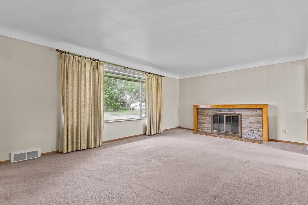 Empty living room with beige carpet, brick fireplace, large window with gold curtains, and crown molding.