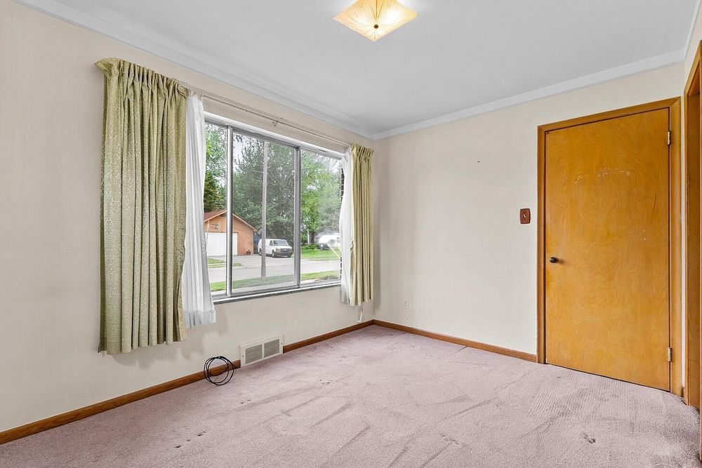Empty bedroom with light pink carpet, crown molding, large window with green curtains, and a wooden door.