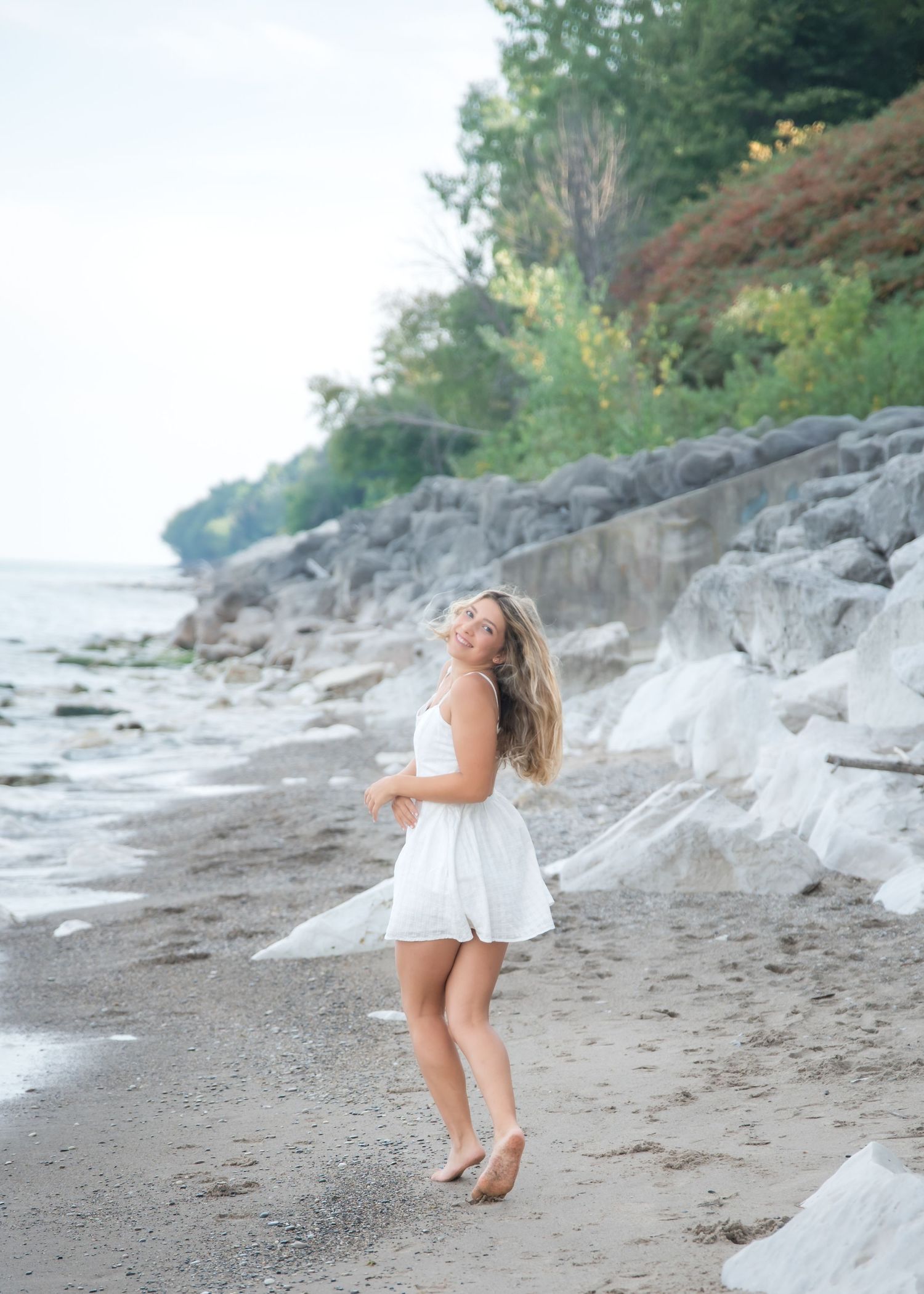 Young woman in white sundress smiling outdoors among rocks and yellow wildflowers on a sunny day.